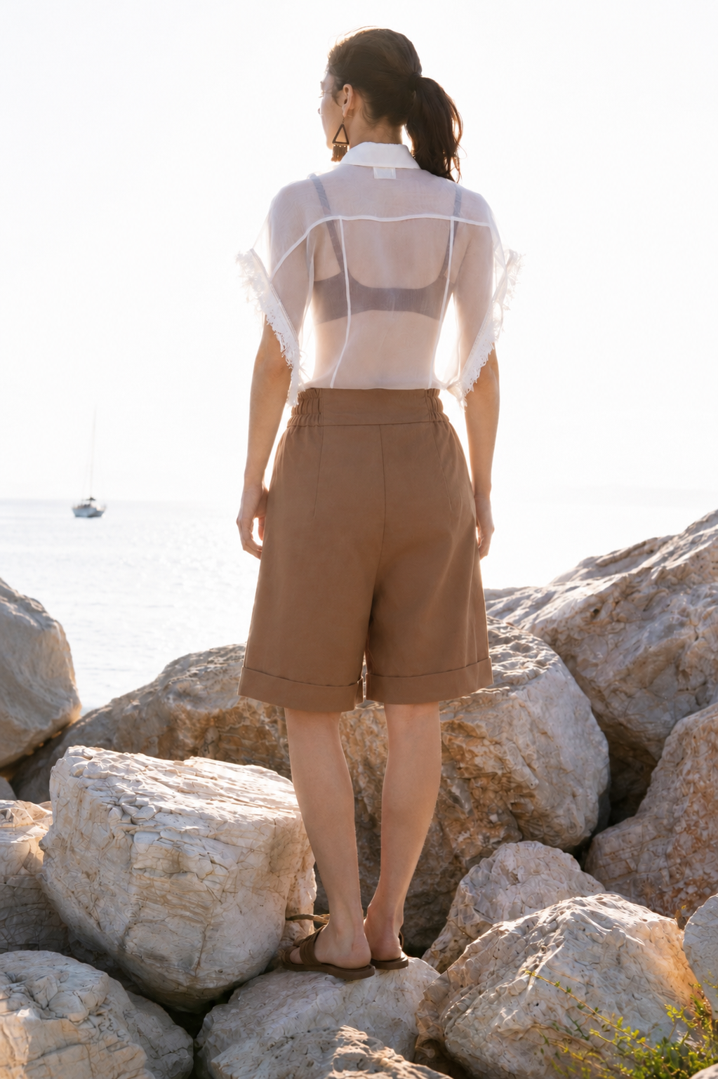 Back view of the Earth Bermuda cotton shorts with relaxed fit and cuffed hem, styled with sheer silk organza shirt on sunlit coastal rocks by Julia Valas