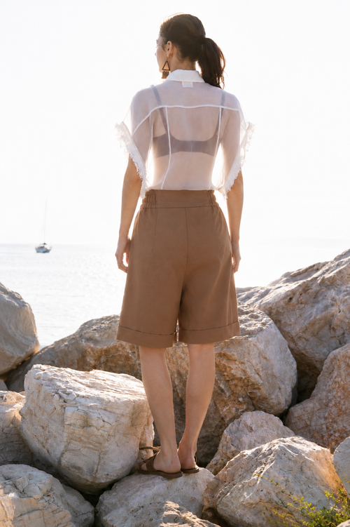 Back view of the Earth Bermuda cotton shorts with relaxed fit and cuffed hem, styled with sheer silk organza shirt on sunlit coastal rocks by Julia Valas