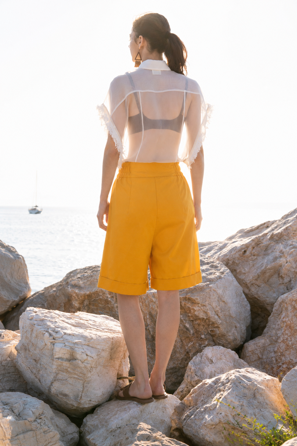 Back view of the Earth Bermuda cotton shorts in canary yellow with relaxed fit and cuffed hem, styled with sheer silk organza shirt on sunlit coastal rocks by Julia Valas