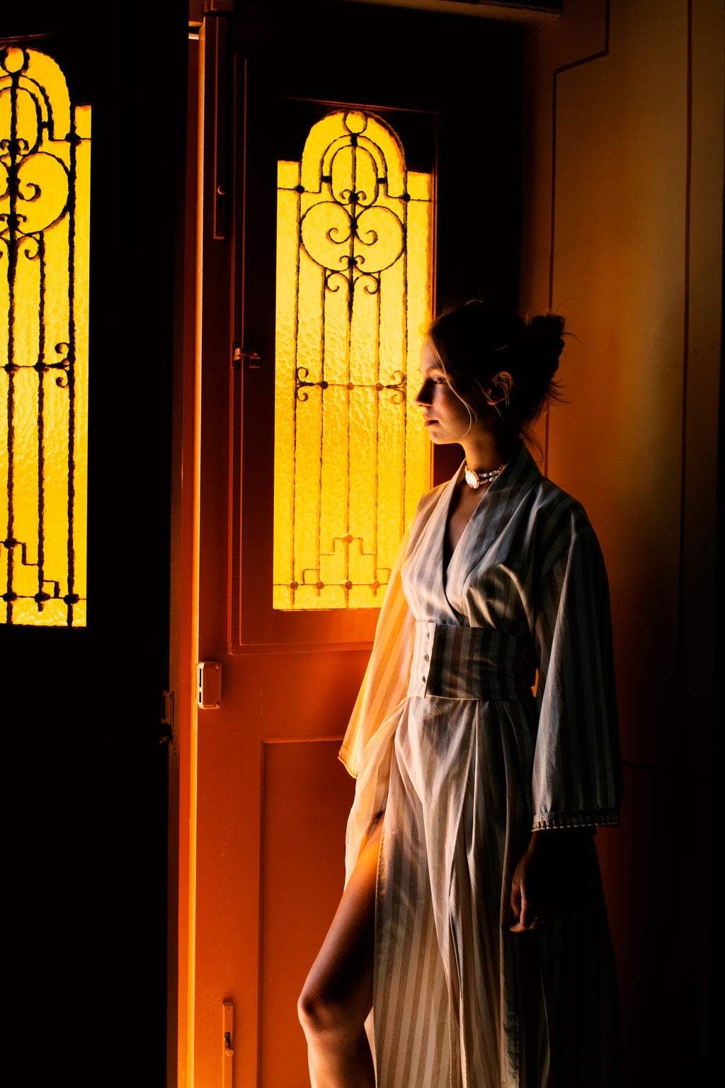 Model wearing the Julia Valas Hour Kimono Dress in Sand Stripe cotton, illuminated by warm light at a doorway in Syros, Greece.