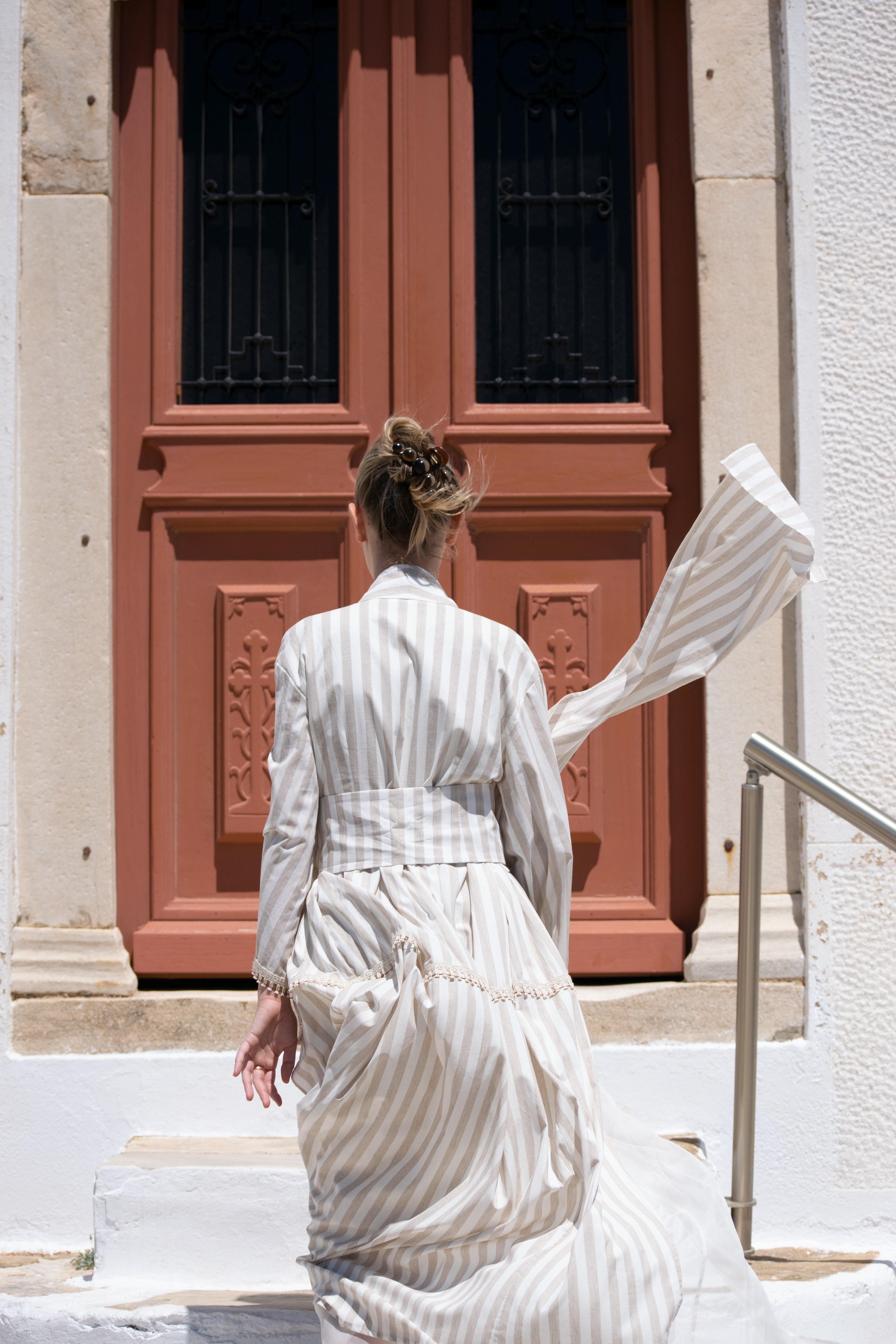 Julia Valas Hour Kimono Dress in Sand Stripe cotton, back view featuring flowing kimono sleeves in the sunlit streets of Syros, Greece.