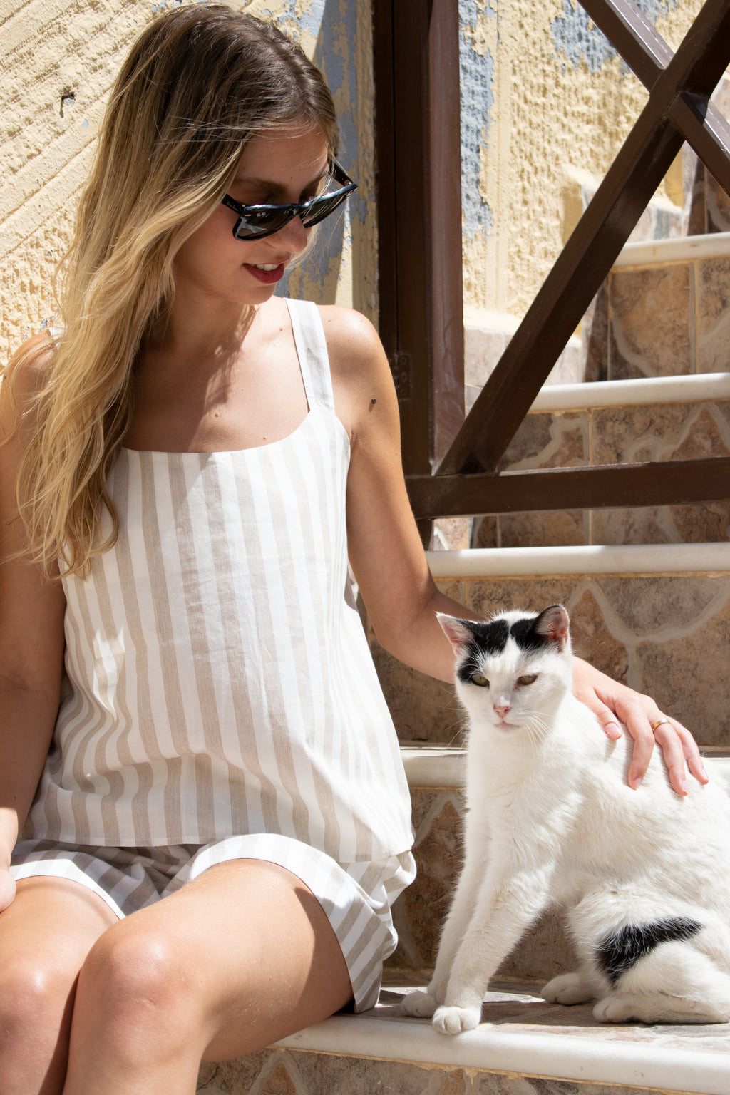 Model wearing the Julia Valas Sleep-to-Seaside Set in Sand Stripe with a striped cotton camisole and relaxed shorts, relaxing with a cat under the Greek summer sun in the Cyclades