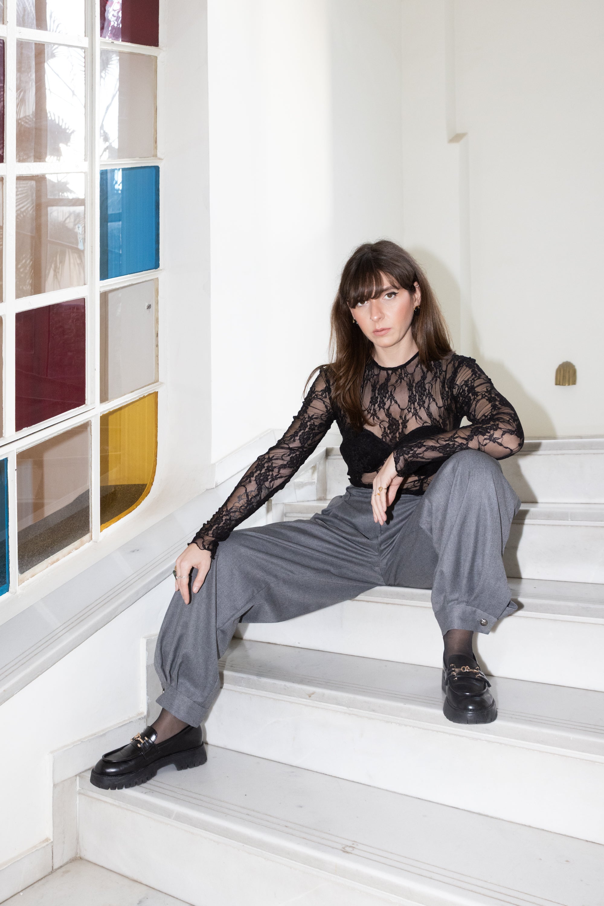 Black lace bodysuit with long sleeves and built-in cups styled with grey relaxed trousers, model sitting on stairs with a wide-leg pose, Julia Valas