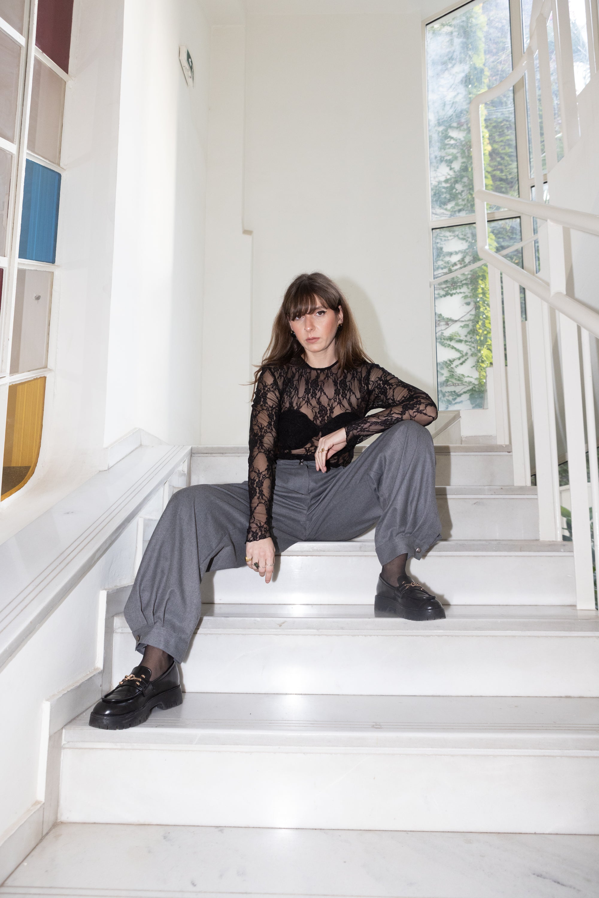 Black lace bodysuit with long sleeves and built-in cups styled with grey relaxed trousers, model sitting on stairs, Julia Valas