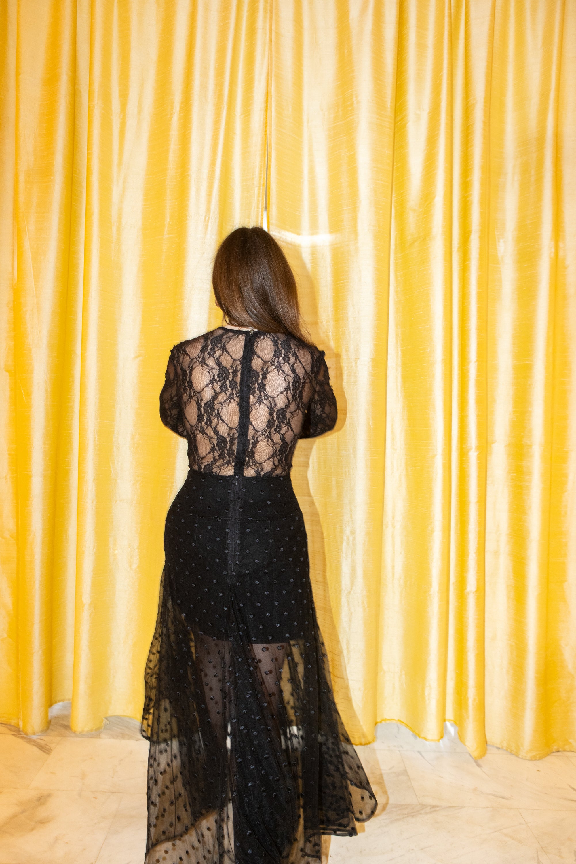 Black lace bodysuit back view with sheer floral pattern and central zipper, styled with flowing polka dot sheer skirt, yellow curtain background, Julia Valas