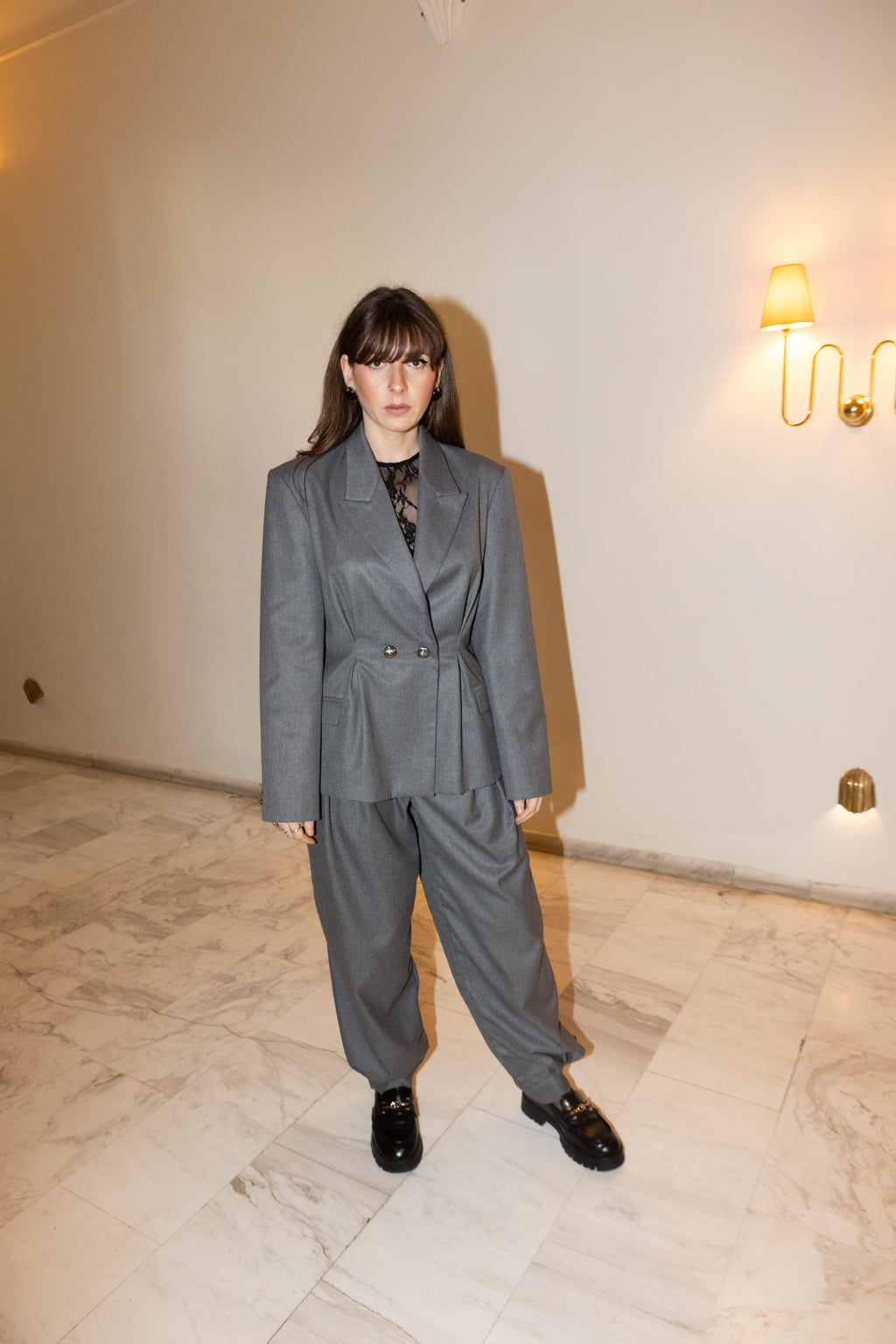 Grey oversized tailored blazer set with pleated waist detail, full body standing pose with slight angle, minimal interior setting, editorial fashion styling