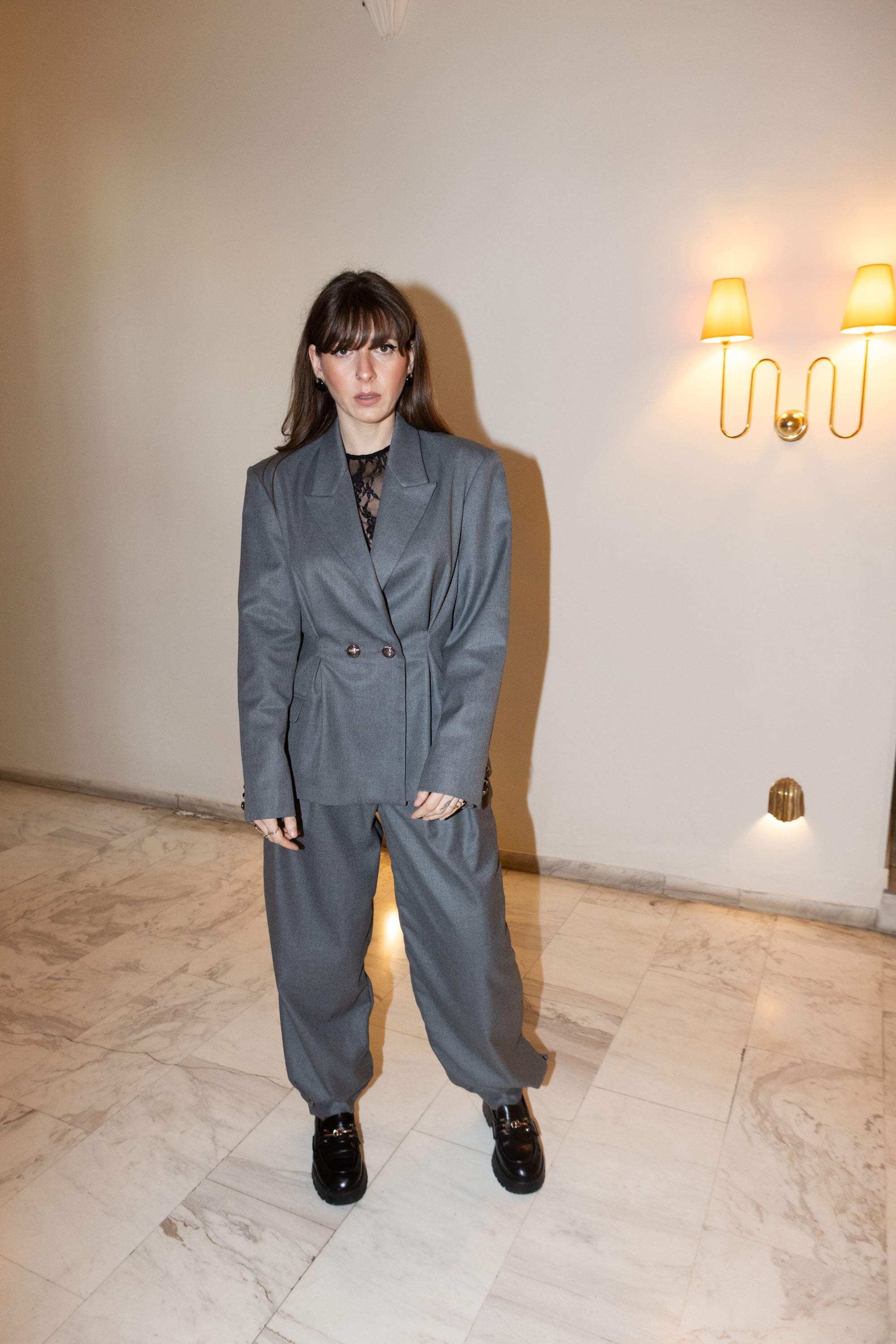 Grey oversized tailored blazer set with pleated waist detail, full body front view, minimal interior setting with soft lighting, editorial fashion styling