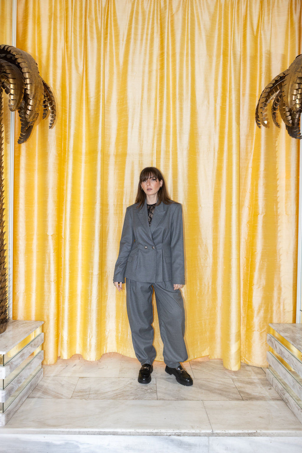 Grey oversized tailored blazer set with pleated waist detail, full body standing pose, gold curtain backdrop, editorial fashion styling