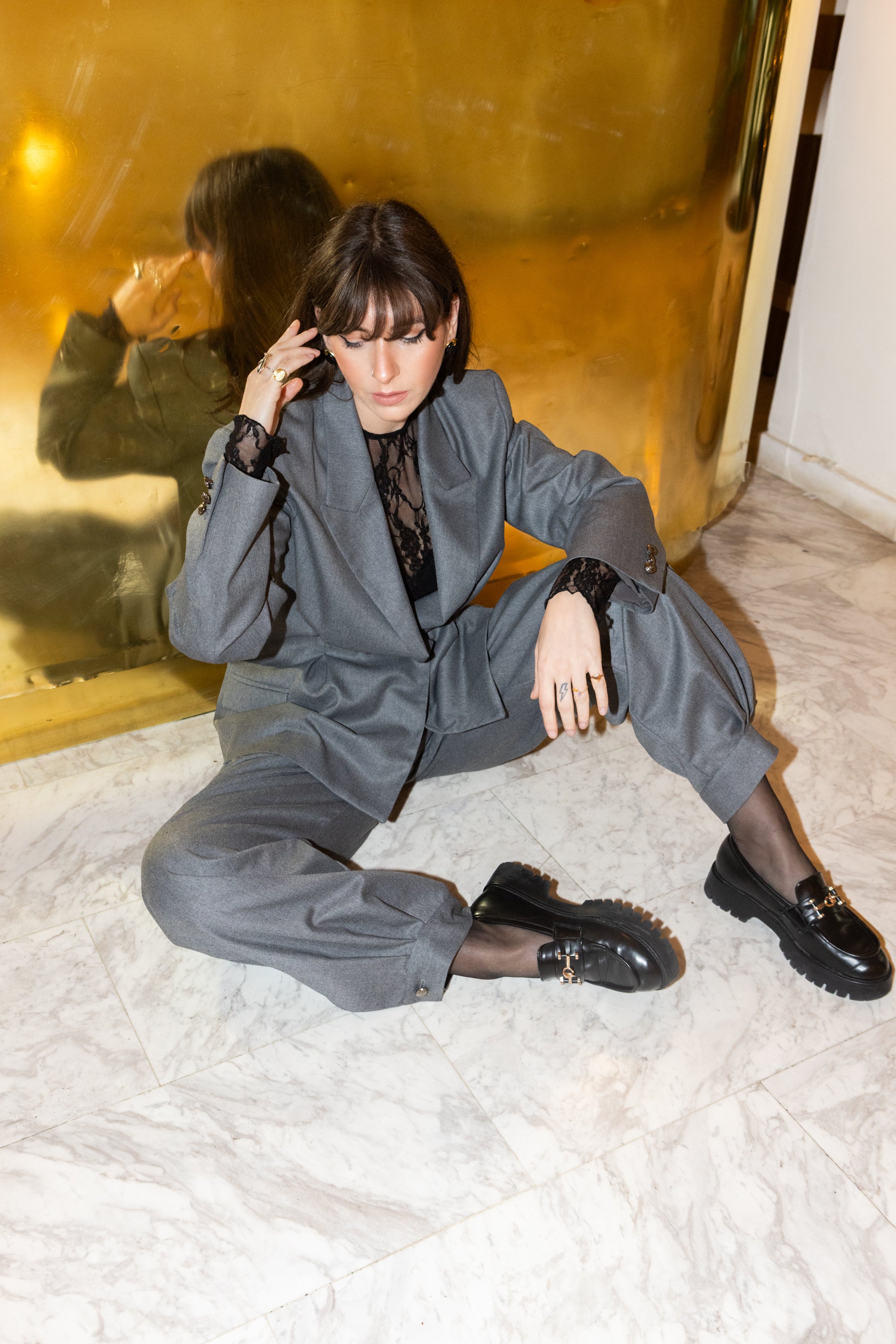Grey oversized tailored blazer set with pleated waist detail, styled with lace bodysuit, sitting pose against gold reflective backdrop, editorial fashion look