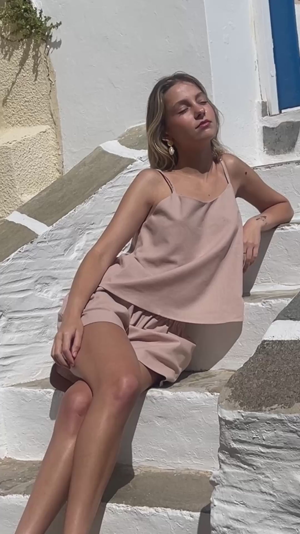 Model wearing Julia Valas Sleep to Seaside Set in Rose Sand featuring a linen camisole top and relaxed shorts and walking in the alleys of a greek island.