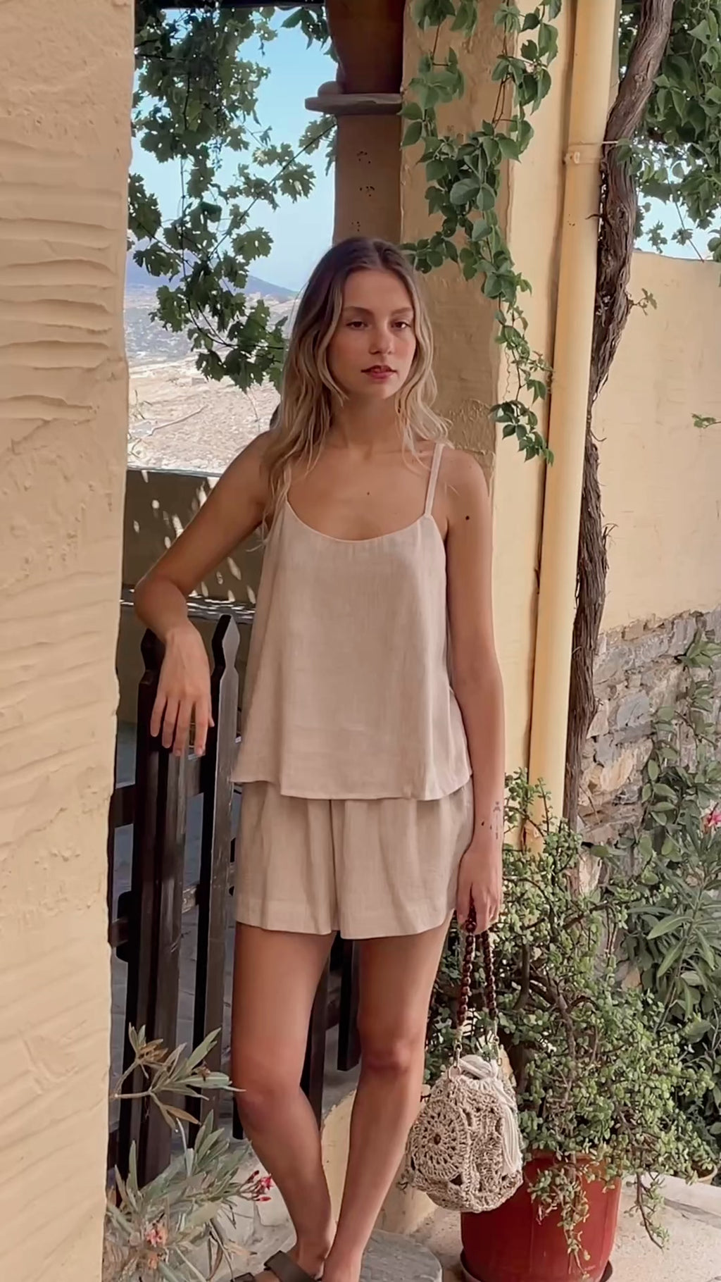 Model walking in cycladic island with the Julia Valas Oat Linen Sleep-to-Seaside Set featuring a sleeveless linen top and matching shorts, standing in a shaded Mediterranean courtyard.
