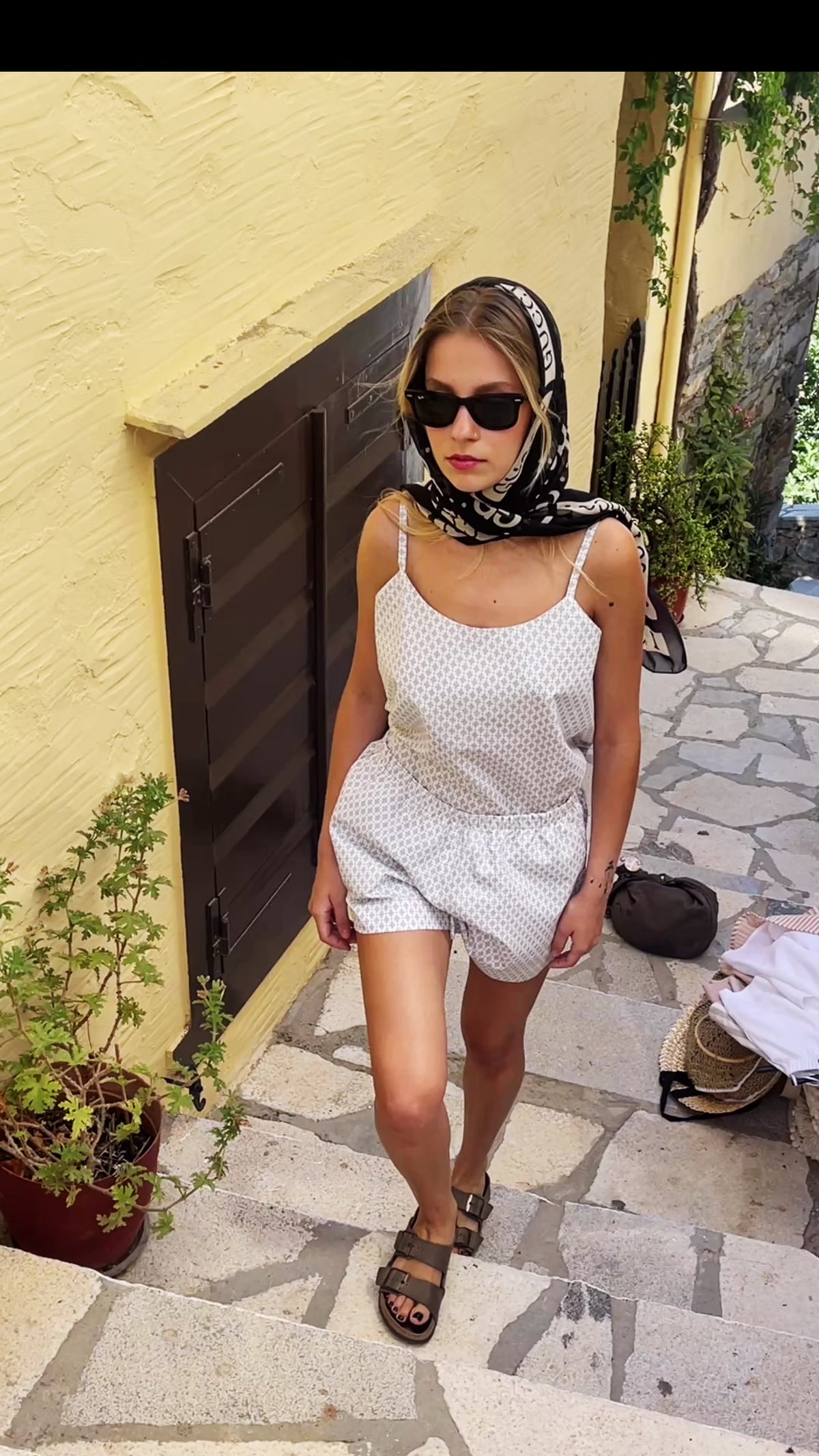Model wearing the Julia Valas Stone Mosaic Sleep-to-Seaside Set walking  in the whitewashed alleyways of the Cyclades, featuring a textured sleeveless top and matching shorts.