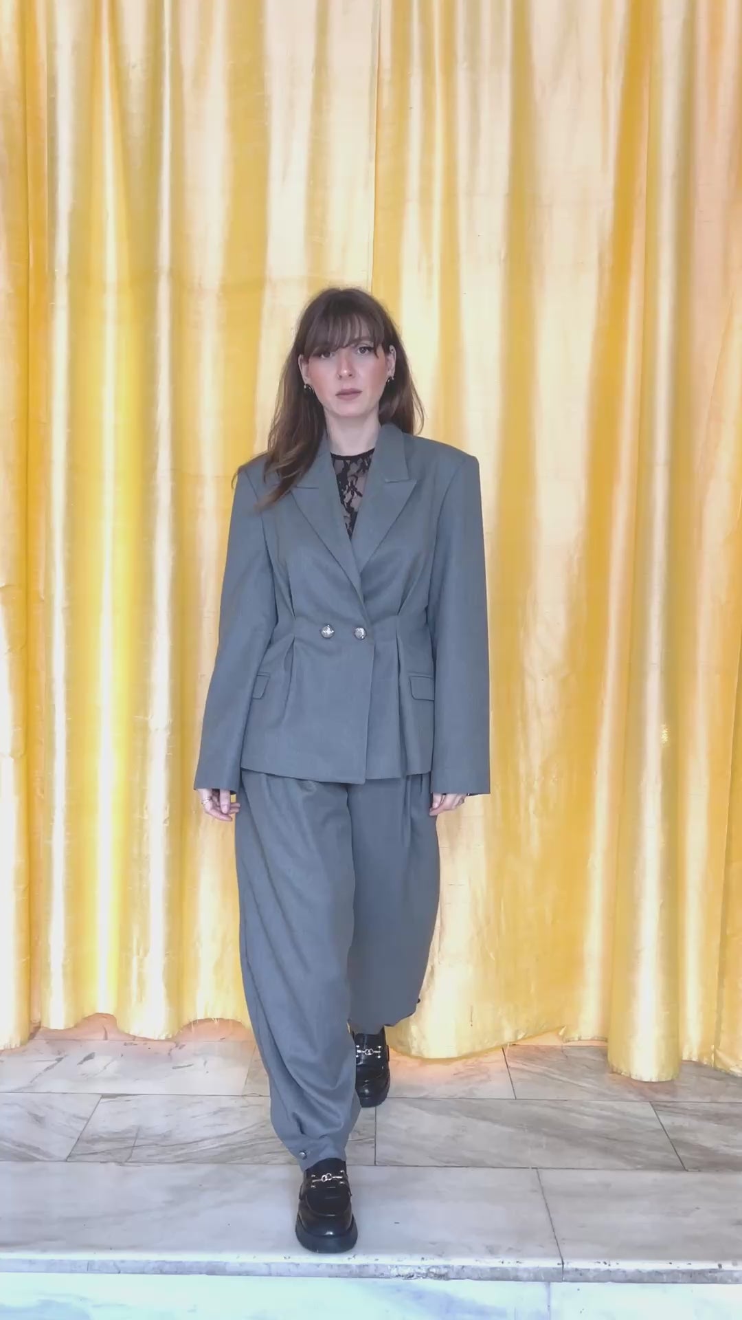 Grey oversized tailored blazer set in motion, model walking forward and back, pleated waist detail, structured shoulders, gold curtain backdrop, editorial fashion video