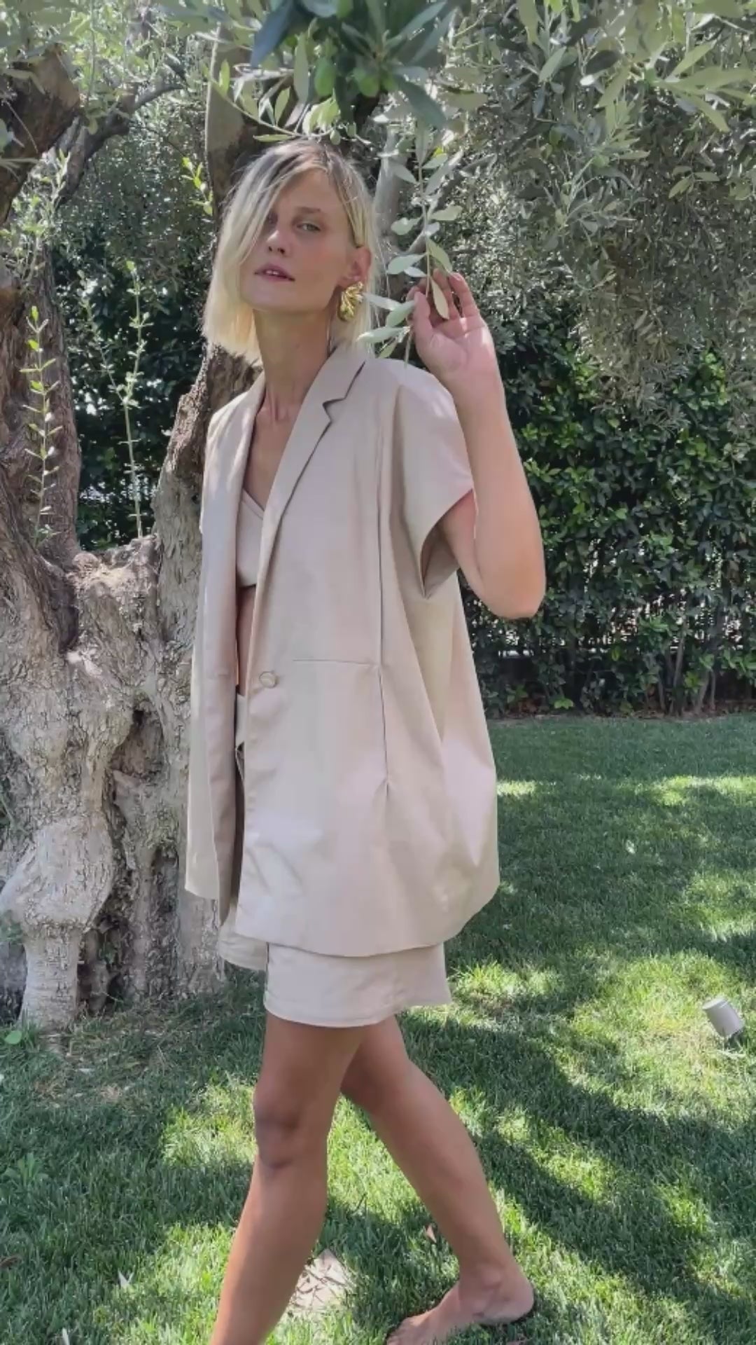 Woman walking under olive tree wearing oversized cotton Halo blazer, bermuda shorts and bralette set