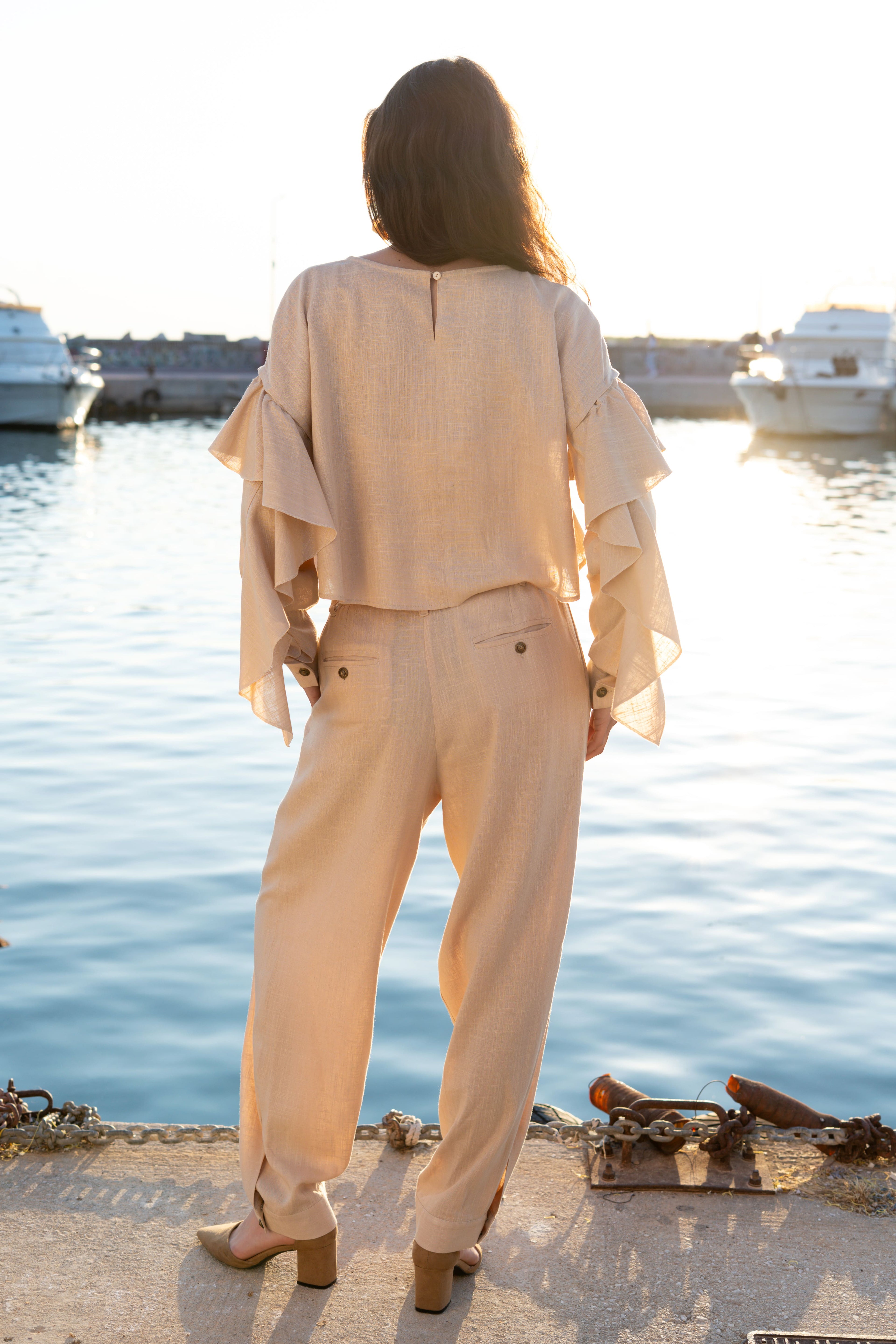 Back view of linen viscose cropped ruffled top with detachable flowing sleeves paired with relaxed pleated trousers, soft minimal resort womenswear by Julia Valas