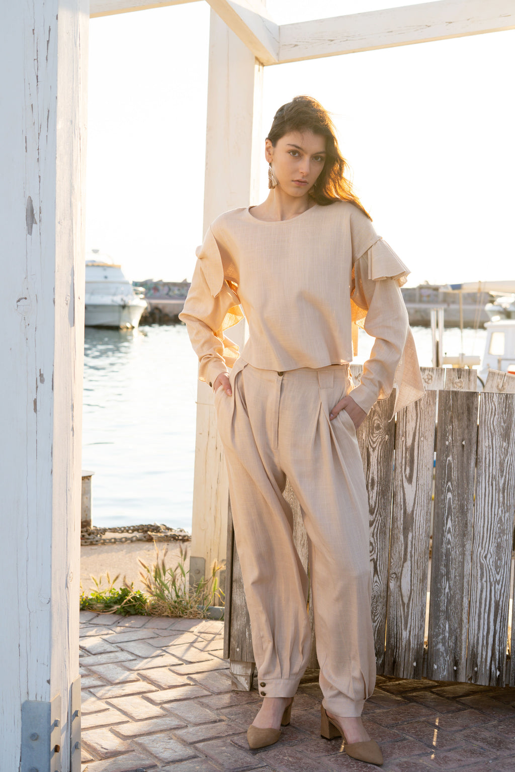 Ruffled linen viscose top with detachable sleeves paired with relaxed pleated tapered pants, soft layered ruffles and elegant summer silhouette in natural sunset light by Julia Valas