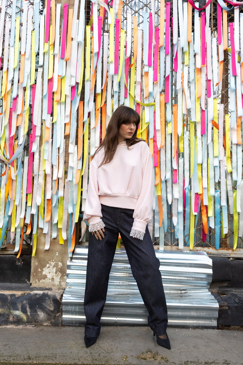 Woman wearing a soft pink cotton lace hoodie with lace cuffs, styled with black trousers, standing in front of colorful ribbons, Julia Valas