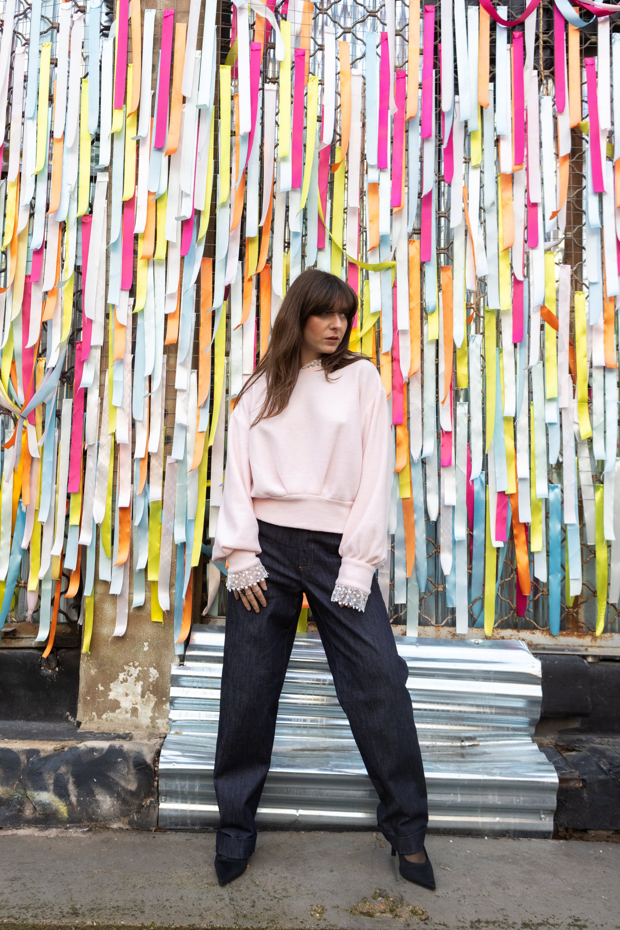 Woman wearing a soft pink cotton lace hoodie with lace cuffs, styled with black trousers, standing in front of colorful ribbons, Julia Valas