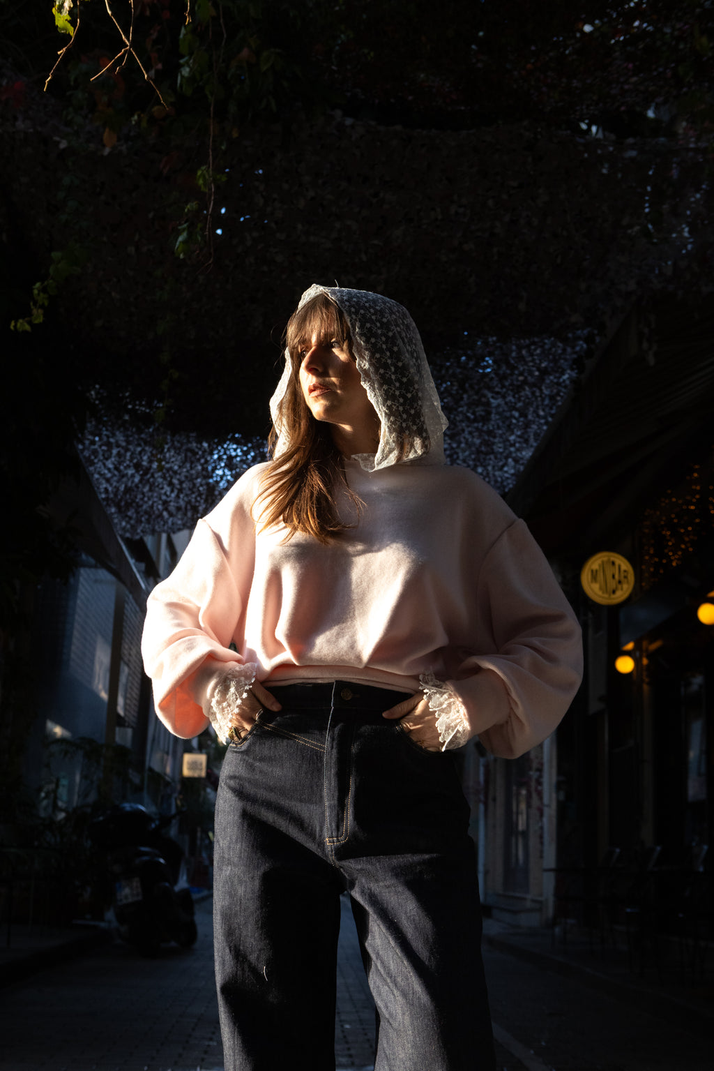 Woman wearing a soft pink cotton lace hoodie with lace hood and cuffs, styled with dark denim, sunlit street setting, Julia Valas