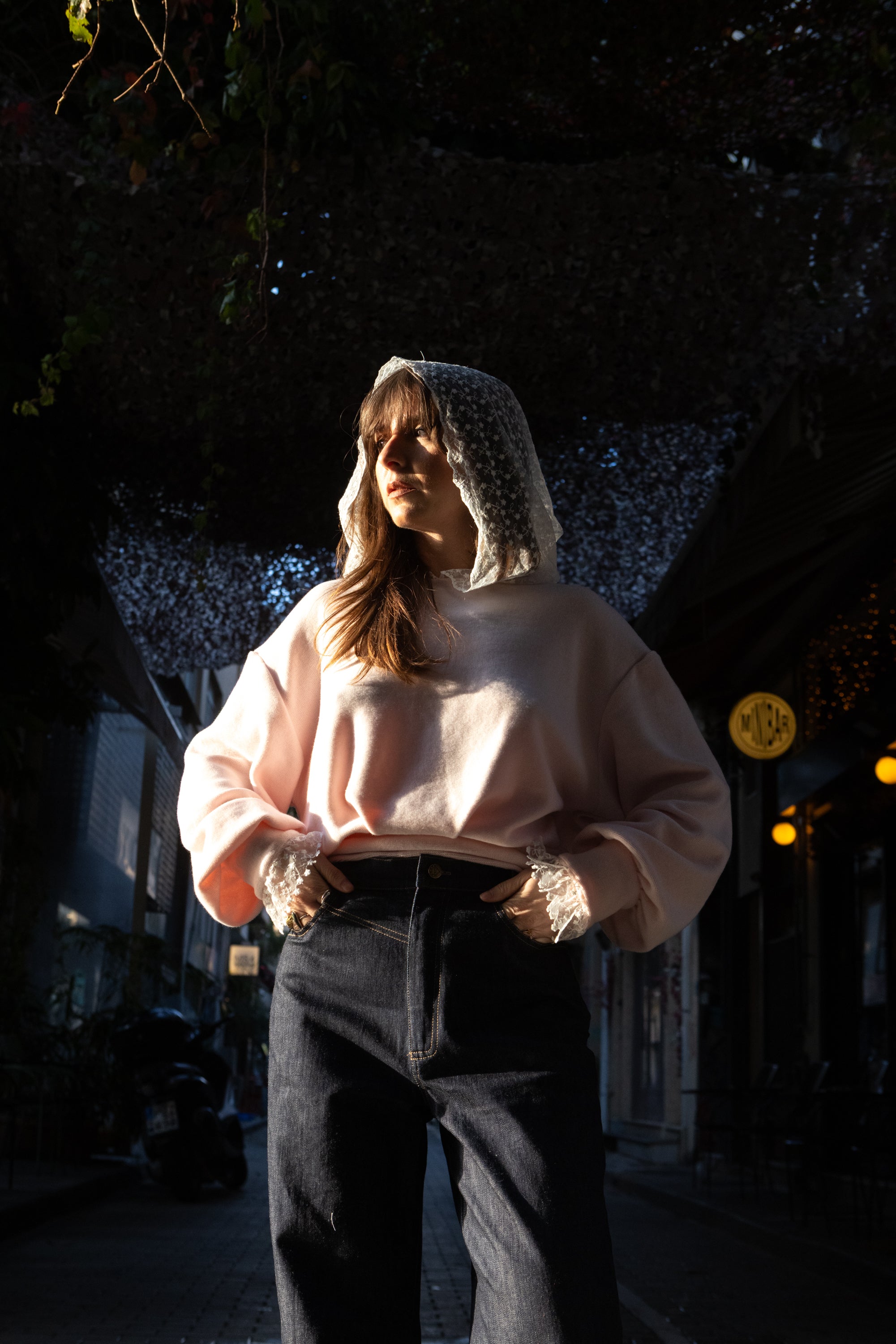 Woman wearing a soft pink cotton lace hoodie with lace hood and cuffs, styled with dark denim, sunlit street setting, Julia Valas