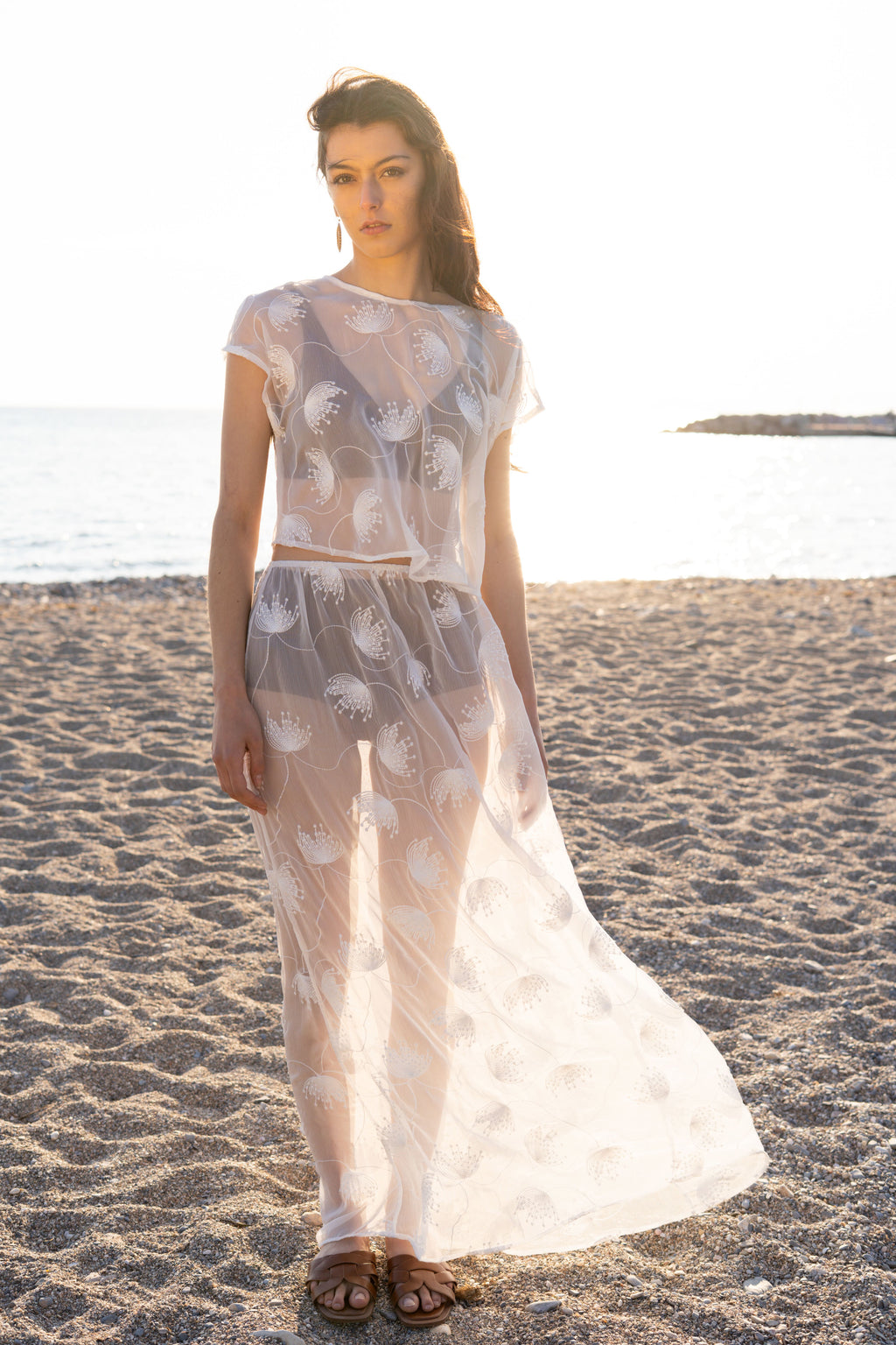 Full-length view of the Thalassa Sheer Set with embroidered transparent top and flowing sheer maxi skirt, lightweight resort silhouette on a sandy beach at sunset by Julia Valas