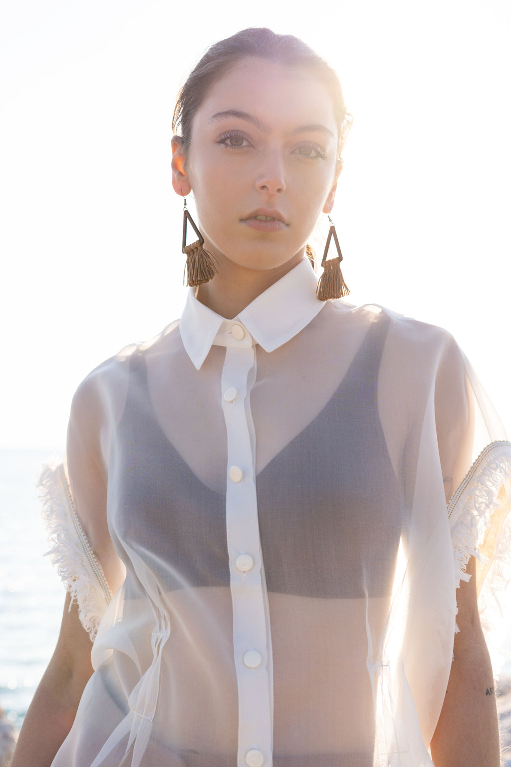 Close-up of sheer silk organza shirt with structured collar, flat buttons and fringed sleeve trim, styled in warm coastal sunset light with visible layered bralette underneath