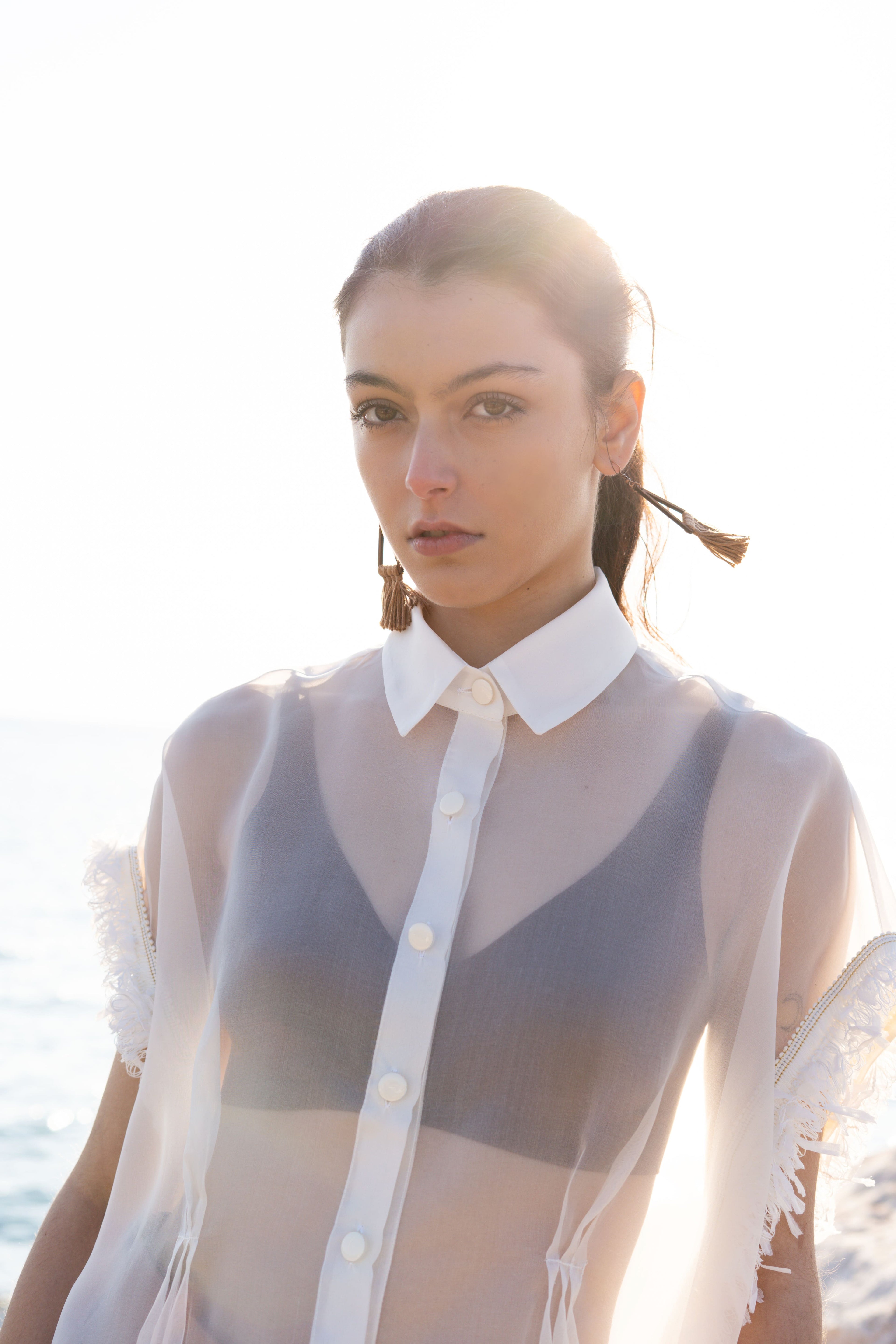 Close-up of sheer silk organza shirt with structured collar, matte flat buttons and fringed sleeve trim, styled over neutral bralette in warm seaside sunset light
