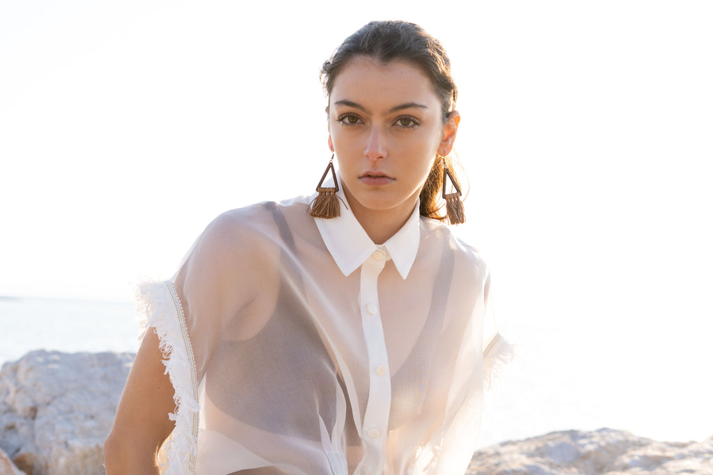 Close-up of sheer silk organza shirt with structured collar, matte flat buttons and fringed trim sleeves, styled over neutral bralette in golden coastal sunset light